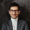 Young, successful businessman wearing glasses, gray jacket, white turtleneck on dark gray background